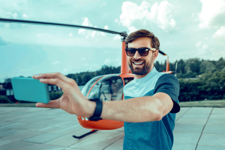 Waiting for flight. Contented short-haired man in stylish outfit making selfie with helicopter on the backgroundの写真素材