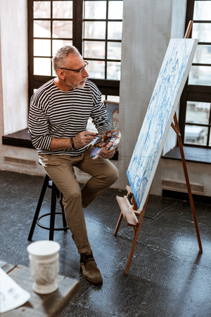 Aged artist. Aged grey-haired artist wearing glasses wearing beige trousers and striped shirt working on new canvasの写真素材