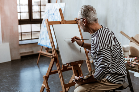 Sitting on chair. Grey-haired experienced artist sitting on chair near painting easel while working hardの写真素材