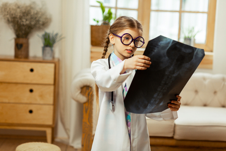 The x-ray in hands. Focused small sweet girl with long brown braids wearing glasses and white coat holding the x-ray in hands.の写真素材