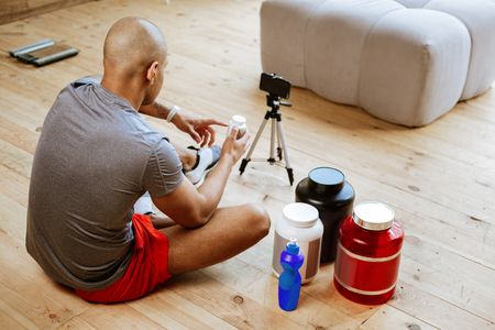 Grey t-shirt. Bald sportsman wearing grey t-shirt sitting on floor and speaking on camera while filming blogの写真素材