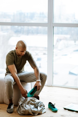 Sneakers into bag. Dark-skinned young sportsman sitting near window and putting sneakers into his sports bagの写真素材