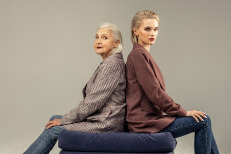 Back to back. Pleasant nice women sitting back to back together on the chair while looking at youの写真素材