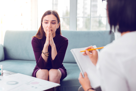 Treatment interview. Professional psychologist carefully making notes during a treatment interview of a brown-haired young-adult appealing depressed sorrowful client.の写真素材