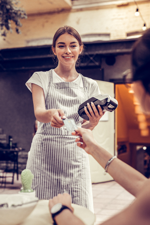 Payment with card. Joyful positive waitress smiling while holding a bank terminalの写真素材