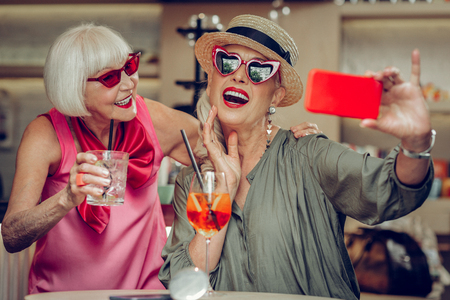 Positive photos. Delighted positive woman holding her smartphone while taking a selfie together with her friendの写真素材