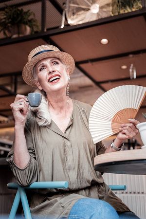 Cooling with a fan. Cheerful fashionable senior lady carrying white fan and cup of strong coffeeの写真素材