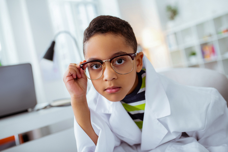 Dark-eyed boy. Cute dark-eyed boy wearing glasses and white jacket of father while visiting him in laboratoryの写真素材