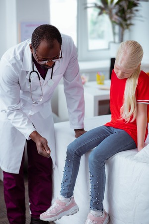Checking knee. Dark-skinned pediatrician in white coat checking knee of blonde stylish girl