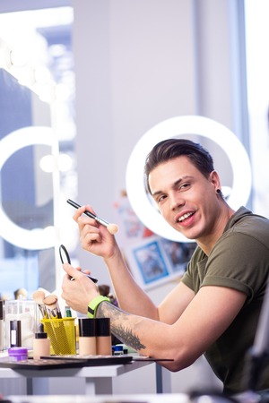 Professional makeup artist. Dark-haired handsome man carrying makeup brush and skin powder while...