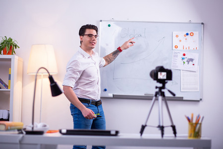 Desk with graphics. Experienced short-haired guy in clear glasses showing desk with tattooed handの写真素材