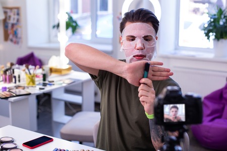 Self-confident young man. Experienced makeup artist sitting covered in moisturizing face mask and presenting interesting product on cameraの写真素材