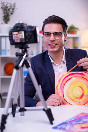 Talking to audience. Smiling communicative young online trainer working with mental issues and showing painting exercisesの写真素材