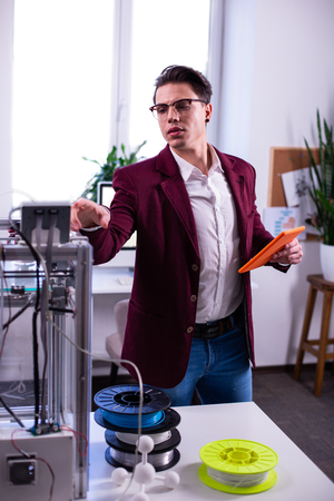 Carrying orange tablet. Professional young engineer wearing blue jeans and checking 3d-printer and...