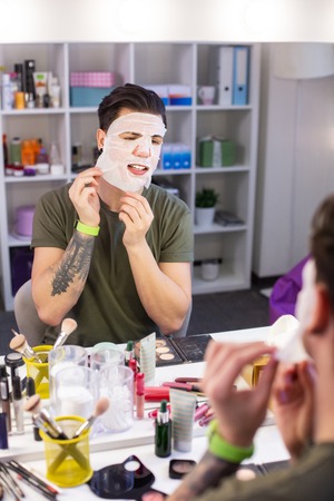 Guy treating skin. Dark-haired guy taking off face tissue mask while sitting in beauty studio and dealing with skin problemsの写真素材