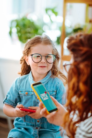Happy daughter. Funny happy daughter looking at mom checking e-mail on smartphone in the office