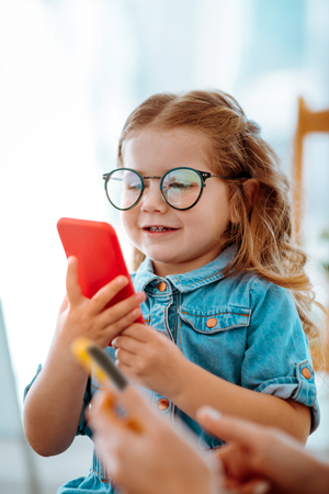 Funny girl. Funny little girl in glasses feeling entertained playing game on phone sitting near momの写真素材