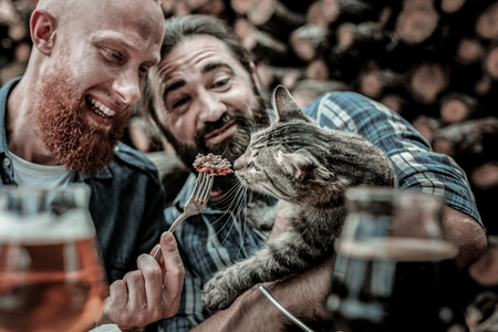Sharing is caring. Two kind smiling friends feeding a stray cat with their burger outside the bar.の写真素材
