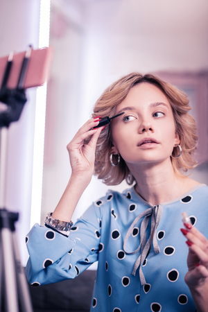 Fixing eyebrows. Blonde blogger fixing her eyebrows with gel while filming video on smartphoneの写真素材