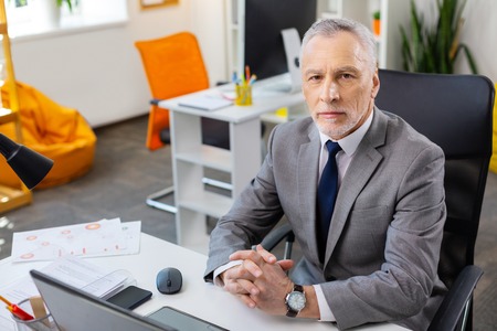 Dealing with laptop. Grey-haired strict man in grey costume being concentrated at work while sitting in black armchairの写真素材
