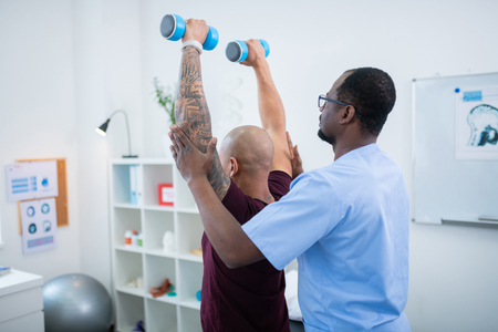 Exercising with barbells. Bald man with tattoo on arm raising hands while exercising with barbells near therapistの写真素材