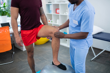 Standing near therapist. Sportsman in red shorts putting leg on fir ball standing near therapist examining his kneeの写真素材