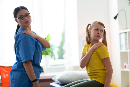 Therapist making posture. Professional physical therapist standing in front of little patient and showing needed posesの写真素材