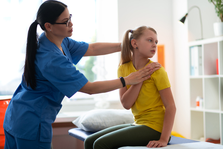 Showing needed poses. Dark-haired doctor in blue uniform presenting special movements while well-behaved patient repeatingの写真素材