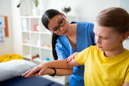 Well-behaved young patient. Attentive Asian long-haired physical doctor in clear glasses examining...