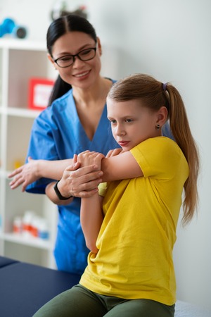 Examining health. Resolute long-haired girl in yellow t-shirt actively stretching hands with support of smiling doctorの写真素材