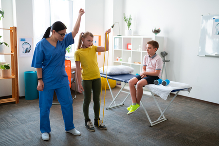 Satisfied with results. Active light haired girl raising hand with yellow resistance rope with instructions of personal therapistの写真素材