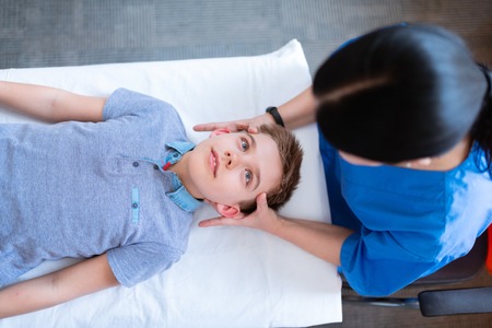 Making head massage. Excited short-haired little boy with blue eyes being relaxed during head massage while lying on the backの写真素材