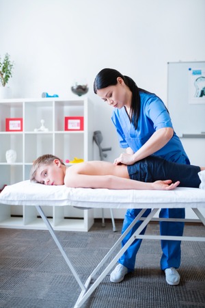 Experienced Asian master. Accurate skilled woman in blue uniform massaging bare back of relaxed little patientの写真素材