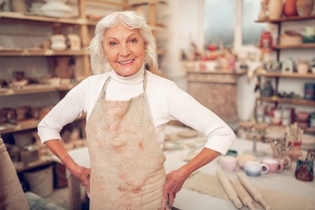 Joyful grey haired woman smiling to you while standing in her workshopの写真素材