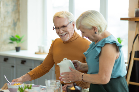 Pouring tea. Kind caring loving old white-haired charming elegant lady in azure top pouring tea for her silver-haired attractive nice-looking spouseの写真素材