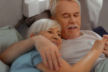 Lying in the bed together. Happy glowing nice-looking loving caring grey-haired married couple in advanced years lying in the bed and hugging each otherの写真素材
