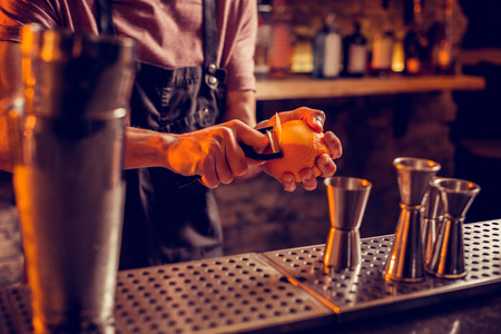Taking orange peel. Close up of professional bartender in black apron taking orange peel for cocktailの写真素材