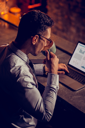 Drinking cappuccino. Elegant businessman drinking cappuccino and working on laptop in his own restaurantの写真素材
