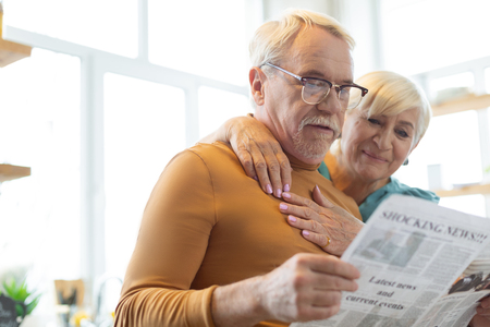 Portrait of cuddling elderly couple. Face portrait of concentrated focused handsome white-haired bearded spouse reading a paper while his gloving cheerful elderly white-haired wife cuddling him.の写真素材
