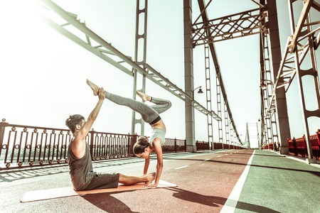 Yoga today. Dark-haired female yogi standing in a handstand pose with her right knee bent while her yoga partner supporting the left legの写真素材
