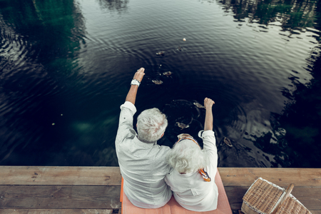 Beautiful scenery. Back shot of nice-looking appealing loving happy elderly spouses with grey short hairdo enjoying the scenery on the lakeの写真素材