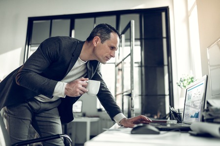 E-mail on laptop. Businessman wearing stylish suit holding cup of coffee reading e-mail on laptopの写真素材