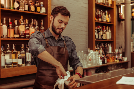 Working at the bar. Handsome attractive dark-haired bearded adult bartender wearing dark clothes and apron polishing glasses at the barの写真素材