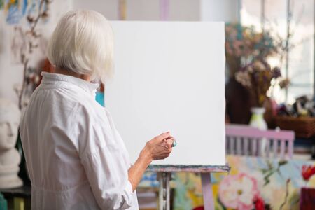 Near white canvas. Blonde-haired aged woman standing near white canvas before paintingの写真素材