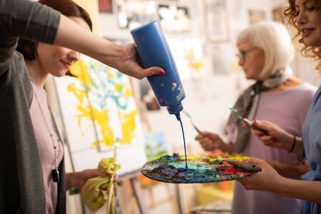 Pouring blue gouache. Dark-haired art student pouring blue gouache on color palette while standing...
