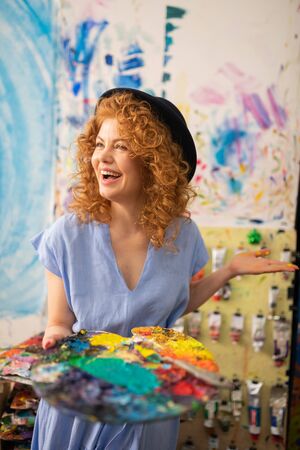 Artist feeling happy. Cheerful curly red-haired artist wearing black hat feeling happy while paintingの写真素材