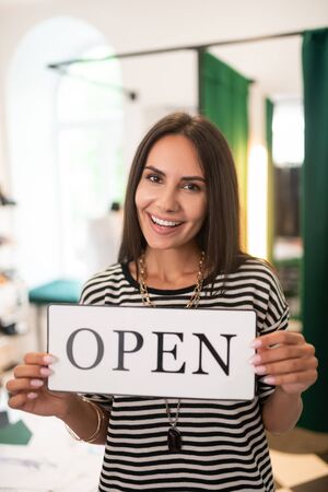 A woman with a sign. Face-portrait of bewitching charming happy smiling cheerful pleasing radiant dark-haired store owner keeping an open sign in handsの写真素材
