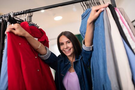Pushing clothes in the store. Close-up portrait of smiling beautiful nice-looking charming dazzling dark-haired female wearing stylish dress and blue cotton jacket pushing clothes in the showroom.の写真素材