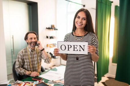 A woman with an open sign. Happy smiling alluring radiant woman holding an open sign while her cheerful joyful attractive partner talking on the phone.の写真素材