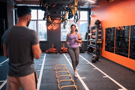 Woman running. Overweight woman wearing grey leggings running in gym smiling to her trainer